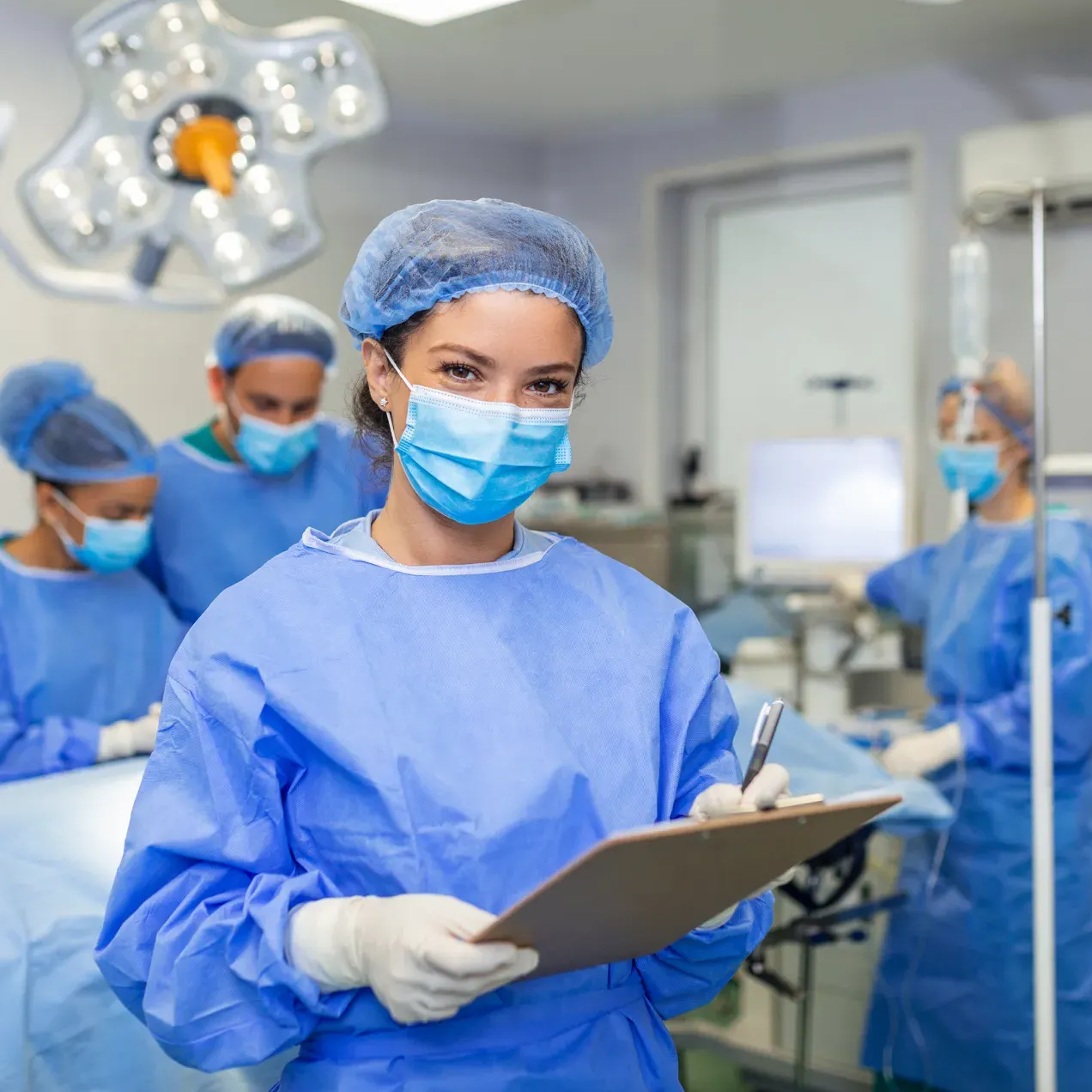 Surgical technology student in scrubs and a mask holding a clipboard in an operating room setting with other students practicing procedures in the background.